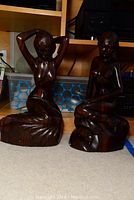 Wide shot showing both dark polished wooden sculptures of sitting nude women side by side on carpeted floor.