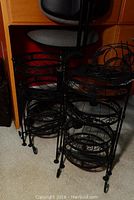 Two black metal four-tiered folding plant stands with scrollwork and wheels shown close together indoors on carpet.