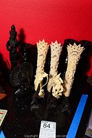 Photo showing 3 carved bone figures with detailed patterns, ebony bust, two ebony bird carvings, and parts of candle sticks against a red background.