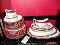 Photo showing stacked plates, gravy boat on saucer, sugar bowl, and other serving pieces with red and gold trim on cream bone china.