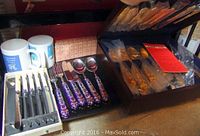 Photo showing cloisonne handled forks, knives, spoons and 2 ceramic mugs with flatware set in box and additional pieces beside.