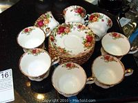 Set of 8 teacups arranged around 8 matching saucers, showing the floral rose pattern with gold accents on a dark surface.