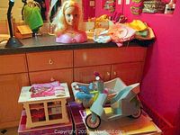 American Girl kitchen counter, bubble bathtub set, scooter, Barbie bust, and doll clothing displayed on kitchen counter in pink room.