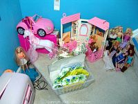 Wide view showing Barbie dolls, pink horse, stable playset, a pink toy car, and basket with doll clothes and accessories.