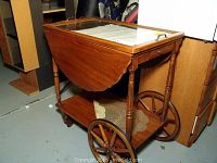 Front angle showing wooden tea cart with glass top, drop leaf side extended, and lower shelf with lace doily.