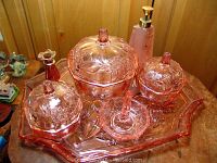 Pink depression glass vanity set arranged on wood table showing tray, lidded dishes, perfume bottle, and ring dish with central holder.
