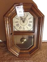Frontal view of Howard Miller clock showing octagonal wooden frame, cream-colored dial with Roman numerals, and glass door with pendulum inside.