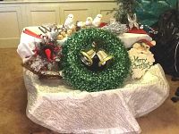 Christmas decorations arranged on a table including a green wreath with gold bells, woodland creature ornaments like a red cardinal, white birds, a small artificial tree, a tree skirt, and a Merry Christmas wall hanging.