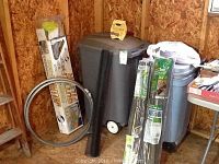 Photo of two large gray wheeled trash cans with lids standing against plywood wall with other items such as metal barrel clamps, rolled screens, and packages leaning nearby.