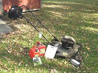 Bolens push lawn mower with handlebar extended, two red gas cans, user manual and accessory packaging laid out on grass with fallen leaves.