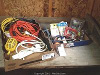 Three outdoor extension cords, a trouble light, surge protector outlet strip, and a box of assorted fasteners and tools arranged on a plywood surface in a shed.
