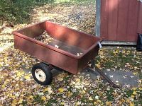 Side angle view of red rectangular metal trailer cart with some rust and dirt leaves inside, sitting outdoors on ground with leaves scattered.
