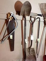 Two hand saws hanging on a wall rack in front with a crowbar, machete, and two garden shovels; all tools show signs of wear and heavy use.