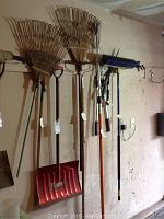 Photo showing three bamboo leaf rakes, one metal garden rake, red plastic snow shovel, push broom, and three pruning shears hanging on wall