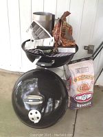 Complete Weber kettle-style grill with charcoal starter, bag of Kingsford Mesquite charcoal, lighter fluid, lighter, and scrubber on a concrete floor near a white wall.