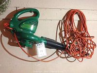 Green handheld electric leaf blower with attached orange electrical cord laid on surface.