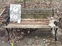 Full view of weathered metal frame wood slat patio bench outdoors with leaves on the ground. Items displayed on bench include the patio clock and fairy statue.