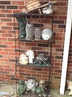 Overview of black metal wire shelf unit with multiple ceramic and resin garden statues arranged on three shelves against brick wall.