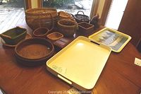 Photo showing collection of 13 assorted woven baskets in different shapes and sizes, two trays including a white plastic tray and a metal painted rectangular tray, and a hardwood carved trinket box on a wooden table.