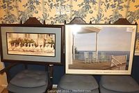 Two framed prints side by side on chairs against a floral wallpaper background. Left print depicts walking ducks, right print depicts a porch with rocking chairs overlooking the sea.