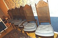 Six wooden dining chairs with cane backs, two of which have armrests. Cushioned seats with additional removable tufted cushions. Chairs have carved crest details on the backrest and curved legs, set together along a wall.