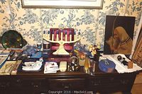 Wide view of collection displayed on wooden sideboard showing menorahs, candles, barware, Judaica figures, books, and original oil painting beside.