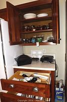 Kitchen cabinet open showing upper shelves with McCoy and Hull brown stoneware dishes, white Pyrex bowls, middle shelves with glassware and small painted dishes. Countertop holds baking trays, small kitchen appliances including Hamilton Beach Mixette and GE mixer, plastic food server and utensils.