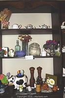 Shelf with multiple glass jars, dried flower arrangements, a white ceramic teapot and other decorative kitchen items including a cow-shaped ceramic figure
