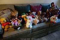 Group of various stuffed animals and dolls arranged on a sofa including Winnie the Pooh, teddy bears, white fluffy dog, green frog stuffed animal, and larger dolls at the right end
