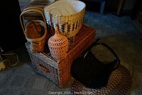Photo showing the wicker trunk, several wicker baskets including a large patterned basket, a small narrow basket, a clay lantern, and part of a woven rug on the floor