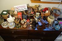 Wide shot displaying the assortment of decorative items on a dark wooden dresser, including baskets, porcelain, glass pieces, candles, and books.