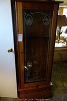 Solid wood vintage gun cabinet with painted glass door featuring wildlife and deer images, two drawers at bottom, locked but no key visible.