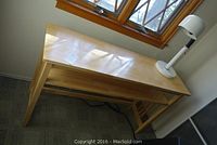 Light-colored solid wood side table seen from front and top angle, showing clean finish, tapered legs, and lower shelf. White metal gooseneck lamp placed on table.