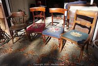 Group of three vintage wooden side chairs with embroidered floral padded seats, placed outdoors showing overall condition and style.