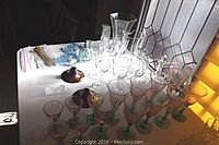 Overview of lot with stemware, spoons, small blue glass dishes, decanter, and vases on a white table near window.