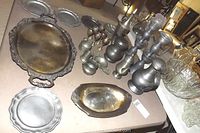 Full view of silver plate serving platters and pewter items on a table including trays, pitchers, candlesticks, and goblets