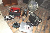 Image shows assorted electronics and camera accessories including a small fan, battery charger, camera, and binoculars.