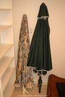 Two closed patio umbrella tops hanging indoors, one solid green with white trim, one with earth-tone geometric pattern.