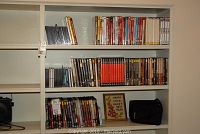 White shelving unit with multiple shelves showing DVDs, CDs, framed cross-stitch and CD binder