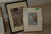 Two framed pictures leaning against a wall; the picture on the right shows an autumn forest stream scene in a brown frame, the other on the left is a darker framed picture with detailed artwork.