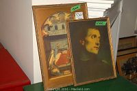 Two vintage framed pictures, one larger with medieval scene and one smaller portrait, on display on a green surface.