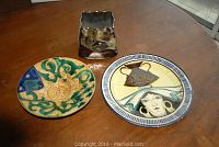 Three pottery items placed on a wooden table: a decorative plate, a bowl, and a square brown vase