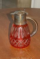 Front angle of cranberry crystal cut glass jug with metal lid and handle on wooden surface.