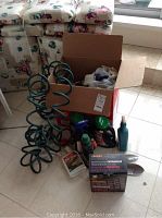 Lot of various garden supplies on floor including coil hose, bottles of garden chemicals, hand tools and a cardboard box with supplies.