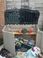 Photo of the open beige plastic garden storage shed with dark green lid showing assorted pots, planters, and gardening tools inside and around it.