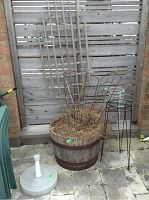 Wide view showing wooden half barrel planter with trellis and dried plants, two metal plant stands beside it and umbrella base on the ground.