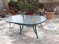 Front view of rectangular glass top table on patio with green metal frame and umbrella hole in center, showing legs and part of umbrella base.