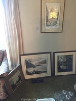 Photo showing three framed art prints arranged near a window and chair: a dark forest silhouette, a river or coastal scene featuring orcas, and another nature scene with orcas. All prints are matted and framed, leaning against wall and floor.