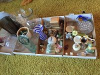 Wide view of three boxes containing assorted glass vases, figurines, ceramic plates, and decorative items arranged on a green carpet