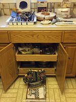 Kitchen utensils including knives, tongs, whisk, spatulas, and measuring cups arranged in a white organizer tray on a kitchen counter with various bakeware items and towels in the cabinet below.